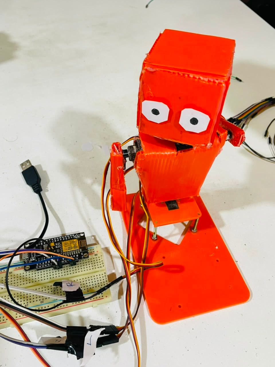 An orange 3D-printed servo robot with two arms, wired to a NodeMCU board